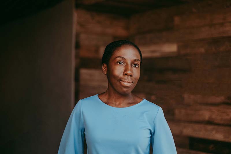 A portrait of writer Jennifer Baker. A black woman, centered in the frame in front of a wooden background, looking directly into the camera with a slight smile. She is wearing a blue shirt.